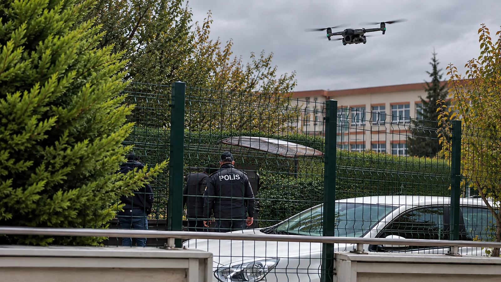 Okul önünde polis ekipleri, güvenlik çiti ve havada uçan drone görülüyor.